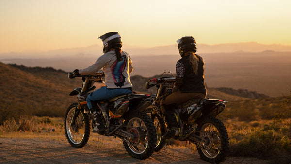 Two women sitting on their dirt bikes looking across the desert at sunset.