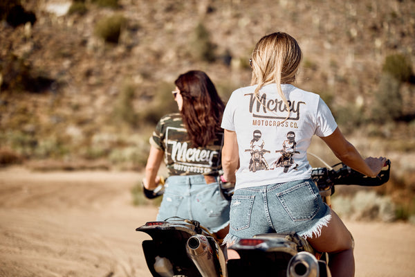 two girls sitting on dirtbikes in the desert wearing short sleeve shirts