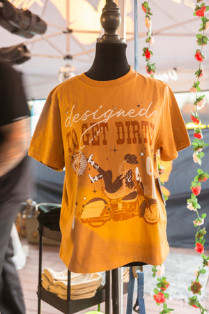 Golden yellow t-shirt with the words designed to get dirty on the front, and a girl sitting backwards on a motorcycle