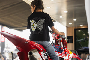 Young girl sitting on a dirt bike, wearing a black shirt with a butterfly on the back. One wing is flowers and the other is black and white checkers.