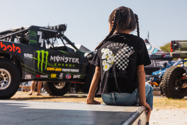 Young girl sitting in front of a Monster Energy truck, wearing a black shirt with a butterfly on the back. One wing is flowers and the other is black and white checkers.