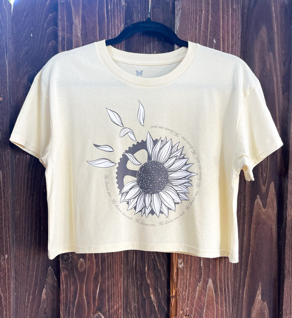 Cropped shirt in butter yellow. There's a sunflower on it with a gear sticking out behind it with the words "He loves me. He loves me not" in a repetitive circle.