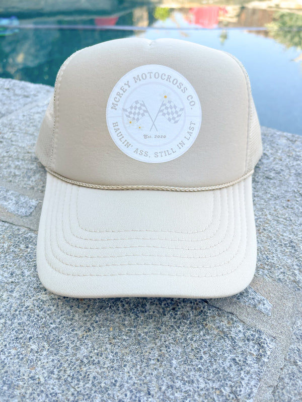 Sand-colored hat with a white circle logo on the front. In the circle it says "MCREY MOTOCROSS CO Haulin Ass, Still in Last" around two checkered flags and three daisies.