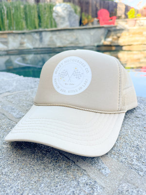 Sand-colored hat with a white circle logo on the front. In the circle it says "MCREY MOTOCROSS CO Haulin Ass, Still in Last" around two checkered flags and three daisies.