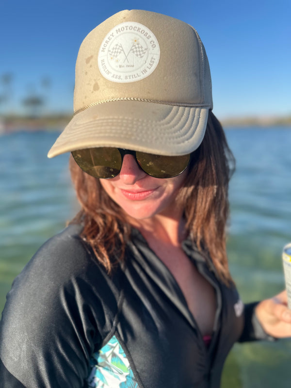 Woman wearing a hat while standing in the river. The hat is sand-colored hat with a white circle logo on the front. In the circle it says "MCREY MOTOCROSS CO Haulin Ass, Still in Last" around two checkered flags and three daisies.