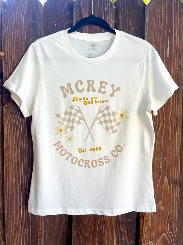 Cream colored 70's-inspired shirt with the words "MCREY MOTOCROSS CO, Haulin' ass, still in last" surrounding two checkered flags and daisies. 