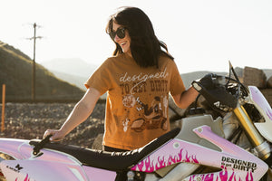Woman wearing a 'Designed to Get Dirty' t-shirt standing next to a dirt bike in an outdoor setting.