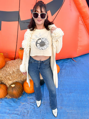 Woman wearing a cropped butter yellow shirt. It has a grey sunflower with petals falling off and a mechanical gear hiding behind it.