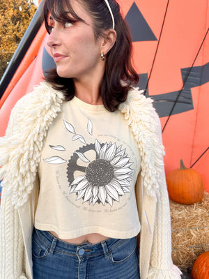 Woman wearing a cropped butter yellow shirt. It has a grey sunflower with petals falling off and a mechanical gear hiding behind it.