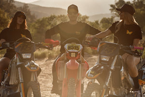 Three friends sitting on dirt bikes, wearing motocross apparel designed for women.