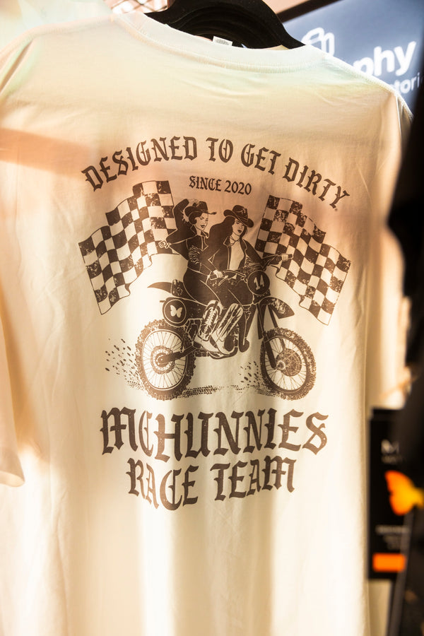 Bone white t-shirt with a graphic on the back showing two girls riding a vintage dirt bike in front of two checkered flags. It says Designed to get dirty since 2020 McHunnies Race Team