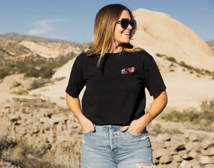 Woman wearing a black t-shirt with a logo and sunglasses, standing in a desert landscape.