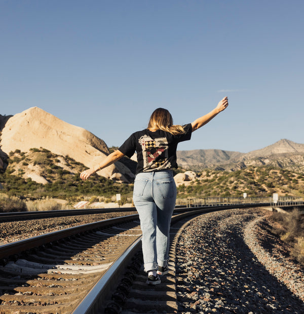 Person standing on train tracks with arms outstretched in a desert landscape