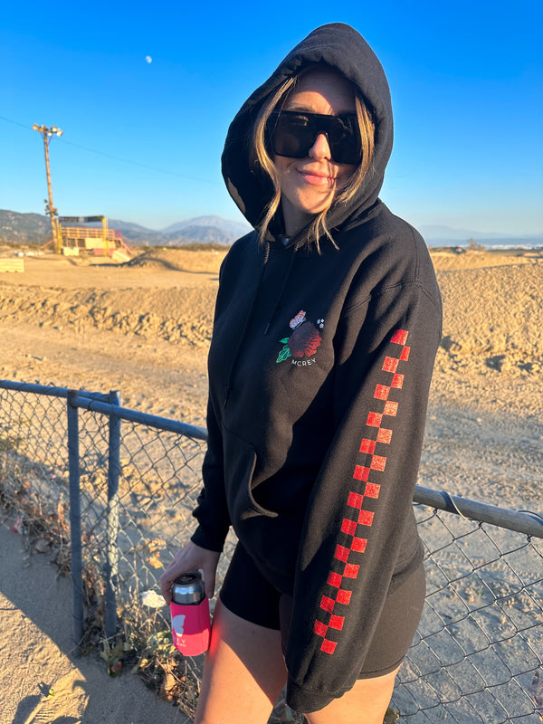 Woman wearing a black hoodie with a flower design on the front and red checkers down the sleeve. She's at a dirt bike track.