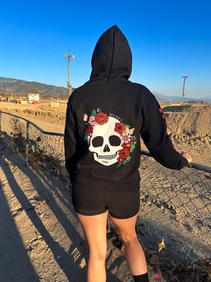 Woman wearing a black hoodie with a skull surrounded by flowers and butterflies on the back. She's watching people ride a dirt bike track.