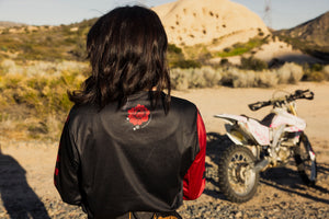 Person wearing a black and red jersey with a flower logo, standing in a desert landscape with an ATV.