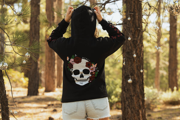 Woman wearing a black hoodie with a skull surrounded by flowers and butterflies on the back. She's watching people ride a dirt bike track.