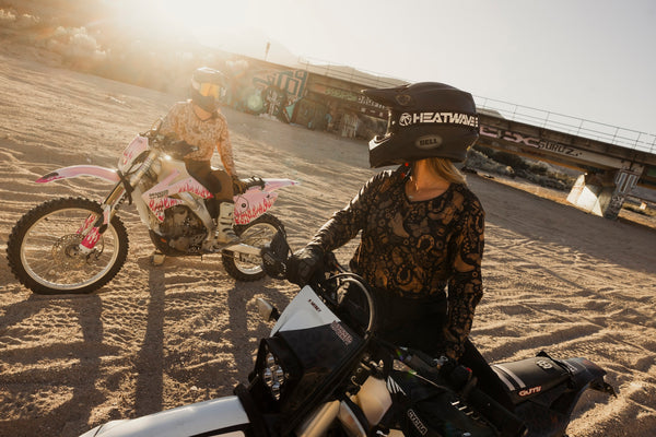 Two women on dirt bikes in a desert setting with a clear sky.