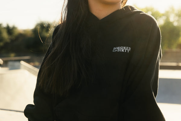 Young girl sitting in a BMX bike park wearing a black hoodie with the words 'Designed to get dirty' on the front