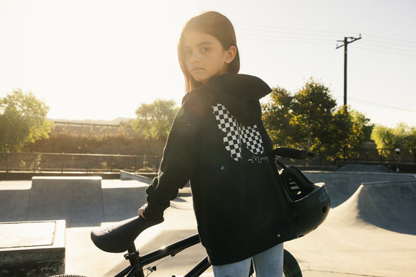 Young girl standing next to a BMX in a bike park wearing a black hoodie with a checkered butterfly on the back