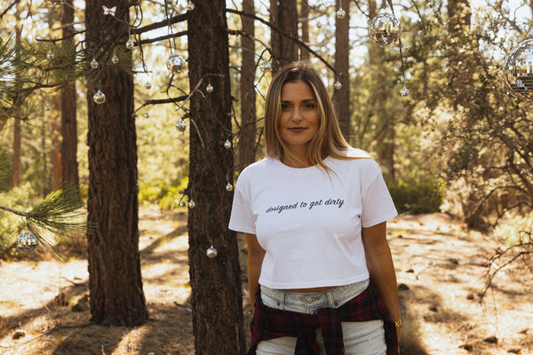 Blonde woman wearing a white crop shirt with black writing that says designed to get dirty standing in front of a tree