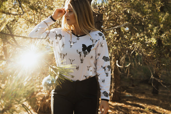 Woman wearing a white dirt bike jersey with grey bull skulls on it. She's standing in the pine trees surrounded by disco balls.