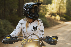 Woman sitting on her dirt bike wearing a white jersey with grey bull skulls on it. She's also wearing a black helmet and black HAVOC goggles