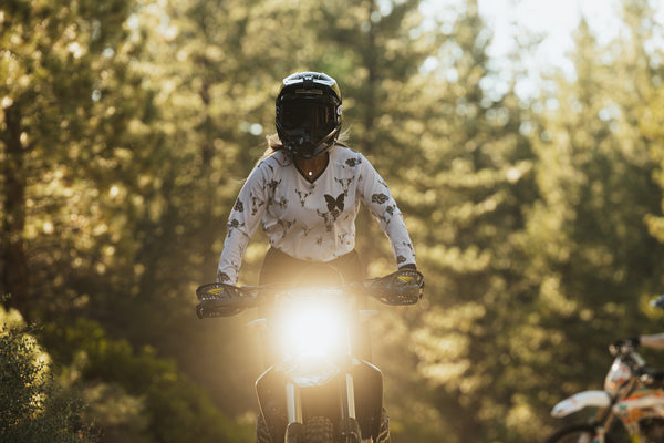 Woman riding her dirt bike wearing a white jersey with grey bull skulls on it. She's also wearing a black helmet and black HAVOC goggles