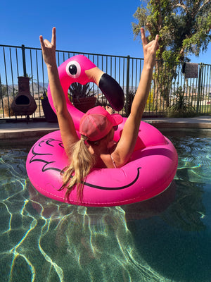 Blonde woman sitting in a pink flamingo in a pool. She is wearing a pink bathing suit and a neon pink trucker hat. She is facing away from the camera with her hands in the air making a rock on sign.