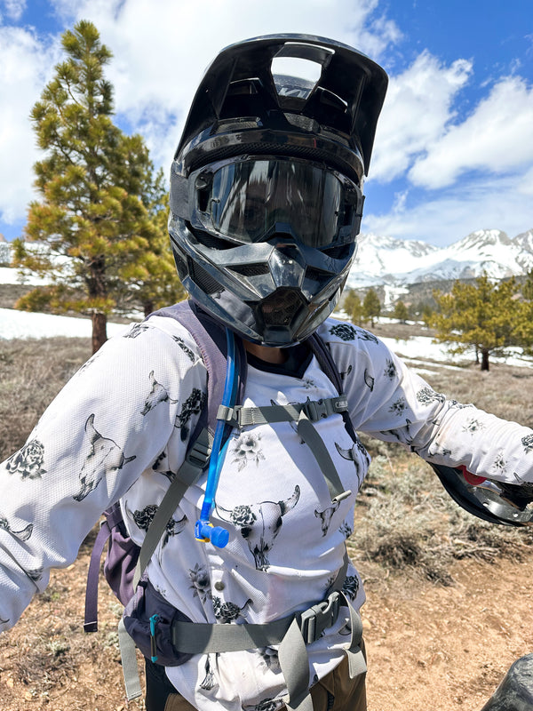 Woman wearing a black dirt bike helmet and white jersey with grey bull skulls on it. She is standing next to her bike in the mountains.
