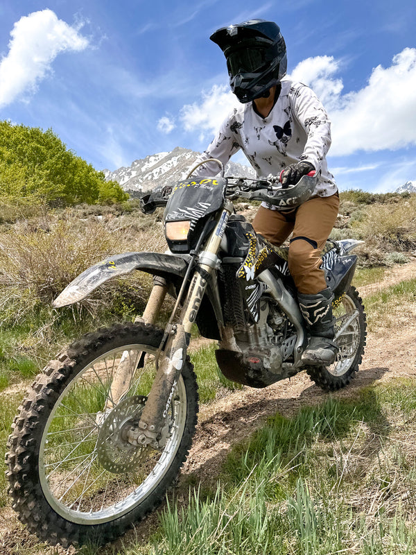 Woman riding a dirt bike down a hill in the mountains. She is wearing a white jersey with grey bull skulls on it and mocha-colored riding pants.