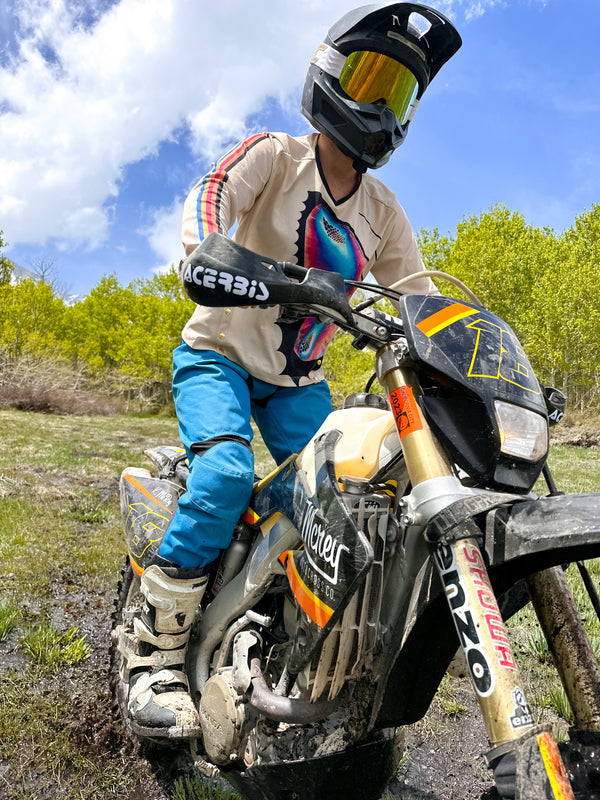 Woman wearing teal dirt bike pants and a colorful butterfly jersey riding her dirt bike in the mud