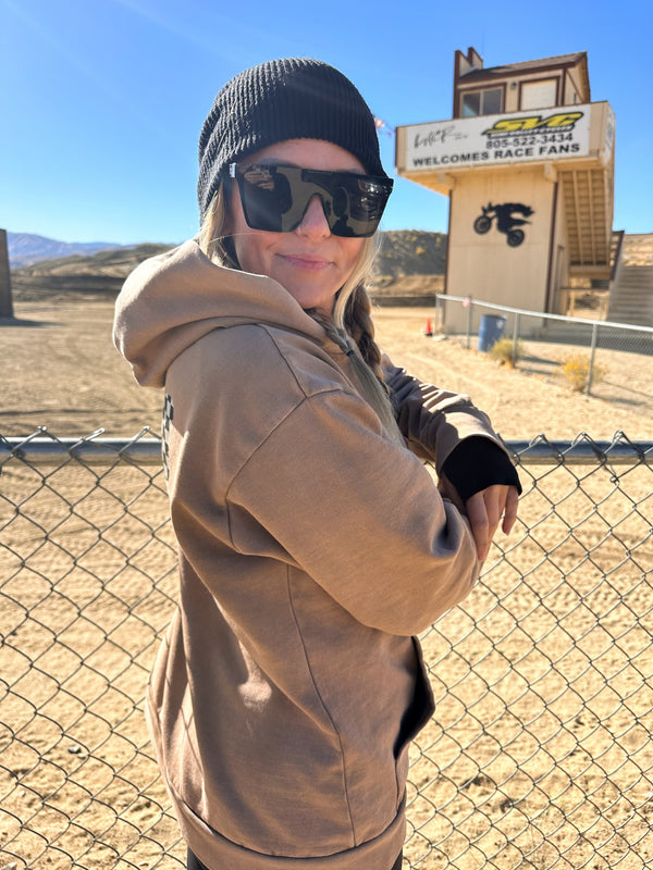 Woman wearing a tan sweatshirt with a black and white checkered butterfly on the back. She's standing in front of a motocross track.