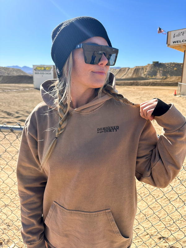 Woman wearing a tan sweatshirt with the words "Designed to get dirty" printed on the front. She's standing in front of a motocross track.