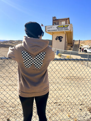 Woman wearing a tan sweatshirt with a black and white checkered butterfly on the back. She's standing in front of a motocross track.