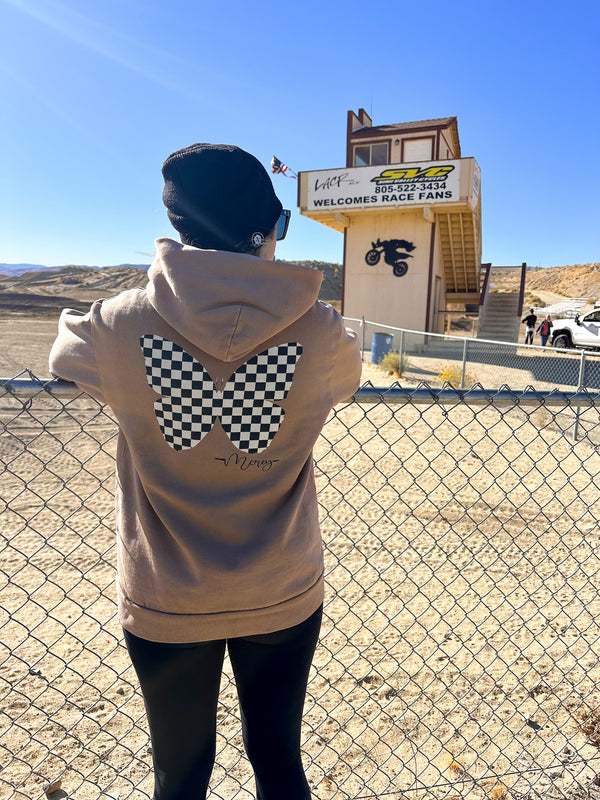 Woman wearing a tan sweatshirt with a black and white checkered butterfly on the back. She's standing in front of a motocross track.