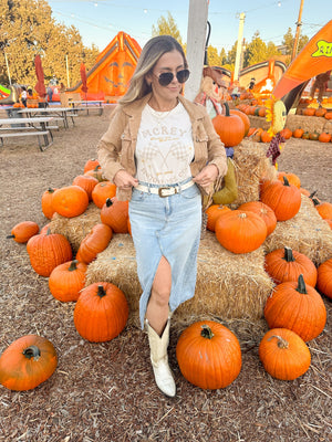 Woman wearing a cream colored shirt with the words 'MCREY MOTOCROSS CO. Haulin ass, still in last' on the front, with checkered flags and daisies. She's standing in a pumpkin patch.