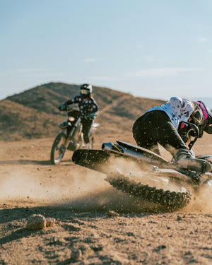 Two women riding dirt bikes. One woman is wearing a white dirt biking jersey and crashing into the ground. The other woman is wearing a navy blue flower dirt biking jersey and riding behind her.