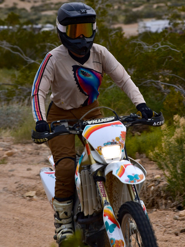 Woman riding her dirt bike wearing a tan jersey with multi-colored stripes down the sleeve and a bright blue and pink butterfly on the front. She is also wearing brown-colored dirt bike pants.