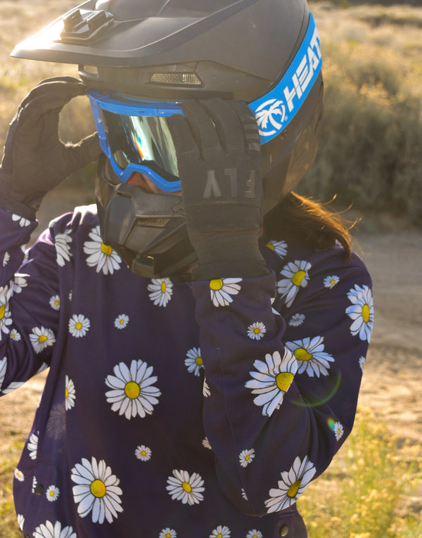 Woman wearing a navy blue dirt bike jersey with white daisies on it. She is putting on blue Heat Wave goggles.