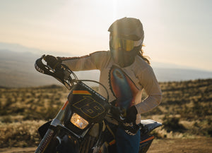 Woman riding her dirt bike in the desert while the sun is setting. She is wearing a tan jersey with multi-colored stripes down the sleeve and a bright blue and pink butterfly on the front.