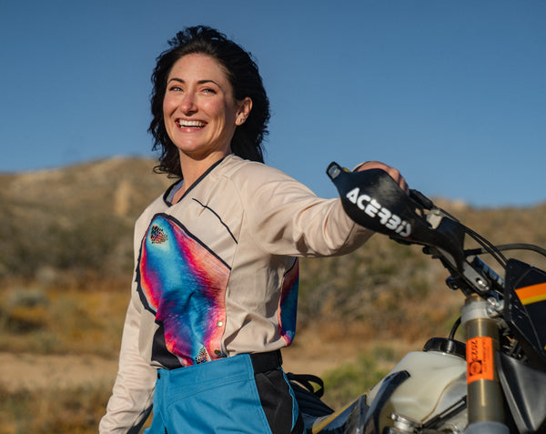 Brunette woman standing in front of her dirt bike, holding onto the throttle. She is wearing a tan jersey with a bright blue and pink butterfly on the front and turquoise pants.