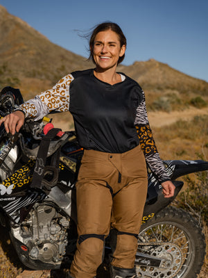 Woman standing in front of her dirt bike smiling into the sun wearing an animal print jersey and mocha riding pants