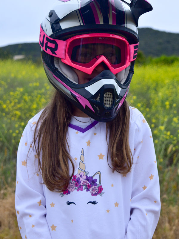 A girl wearing a white dirt bike jersey with a unicorn on the front with a pink and purple flower crown surrounded by stars. 