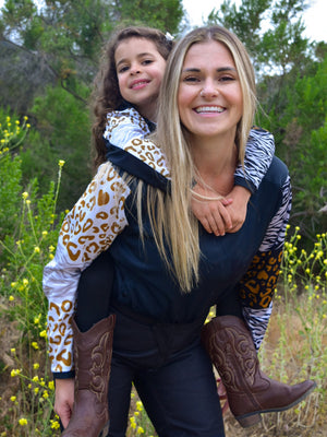 A blonde woman giving a little girl a piggyback ride. They are wearing matching animal print dirt bike jerseys.