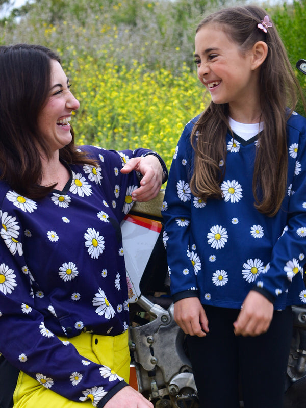 A woman and a girl laughing at each other. They are wearing matching dirt bike jerseys that are navy blue with white daisies on them.