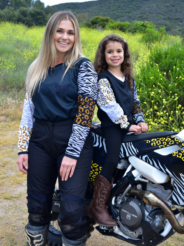 A woman and a girl wearing matching animal print jerseys while the young girl sits on a dirt bike.