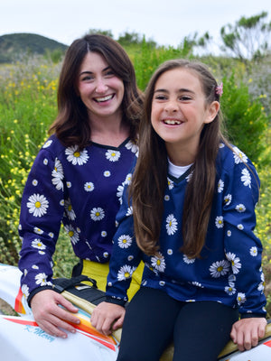 A woman and a girl laughing at each other. They are wearing matching dirt bike jerseys that are navy blue with white daisies on them.