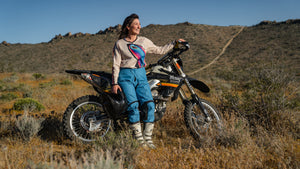 Woman wearing teal dirt bike pants and a colorful butterfly jersey standing in front of her dirt bike