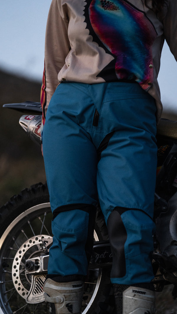 Woman wearing teal dirt bike pants and a colorful butterfly jersey standing in front of her dirt bike